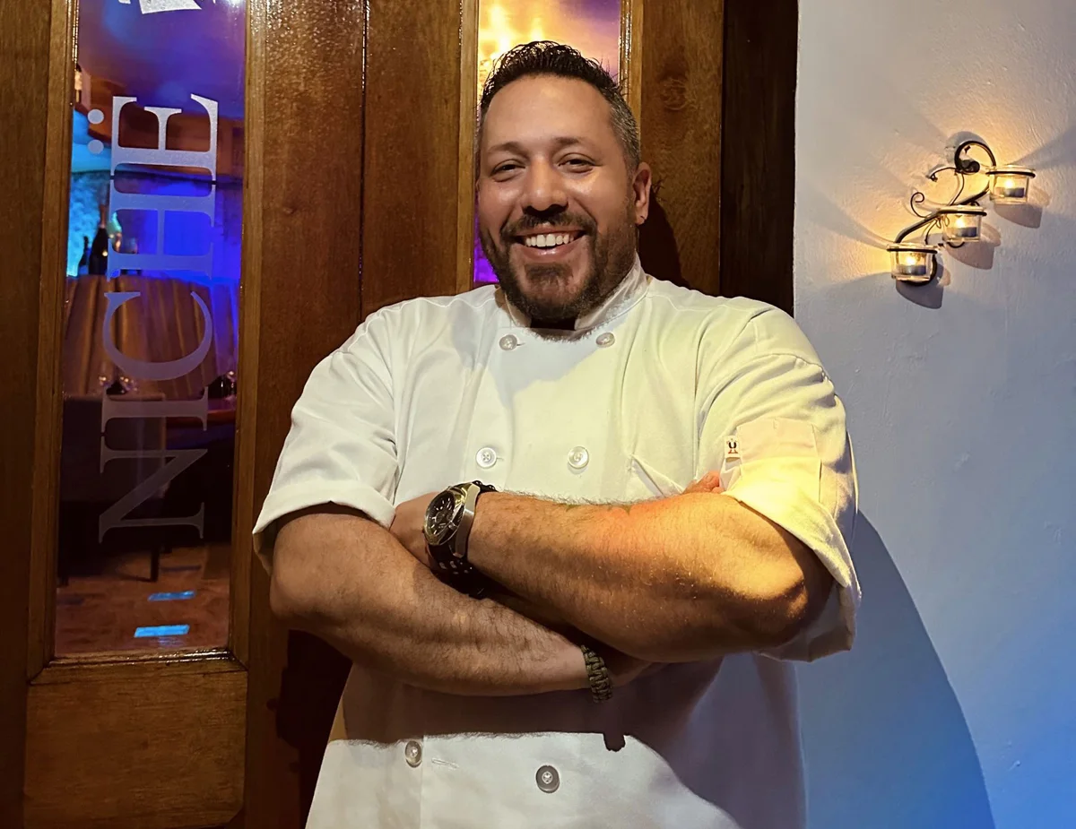 Chef Carlos Morales smiling in chef whites beside the etched-glass Niche door